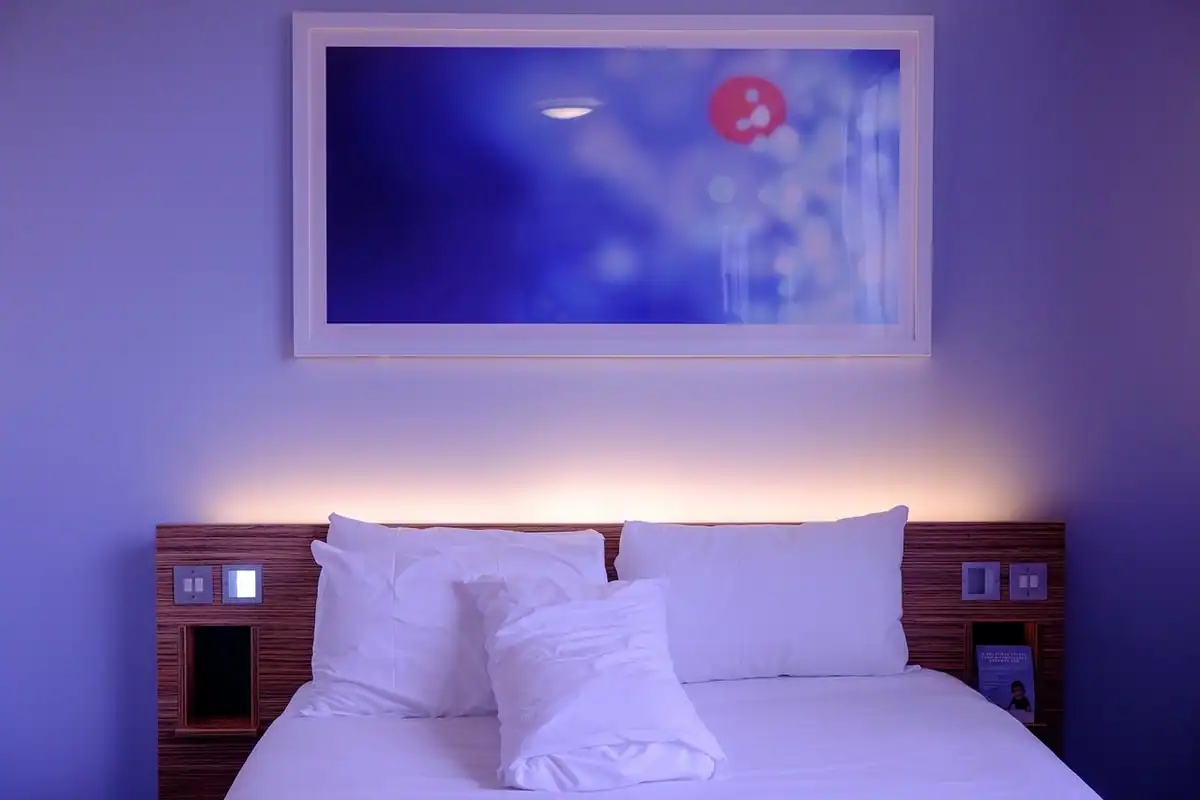Cameră de hotel luminoasă, cu pat mare, lenjerie albă, perne și decor simplu pe perete