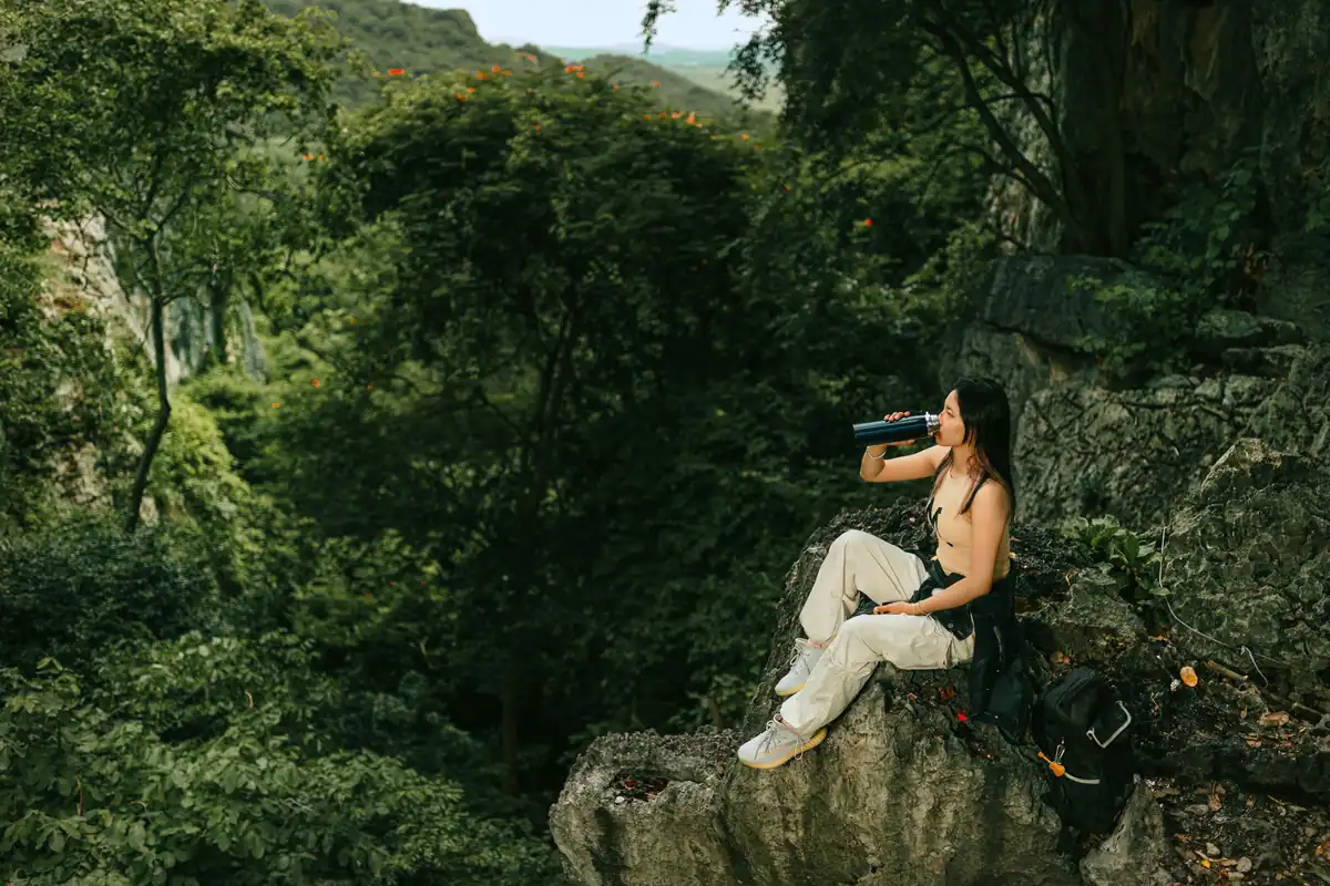 Femeie așezată pe o stâncă, bând apă într-o pădure verde și luxuriantă