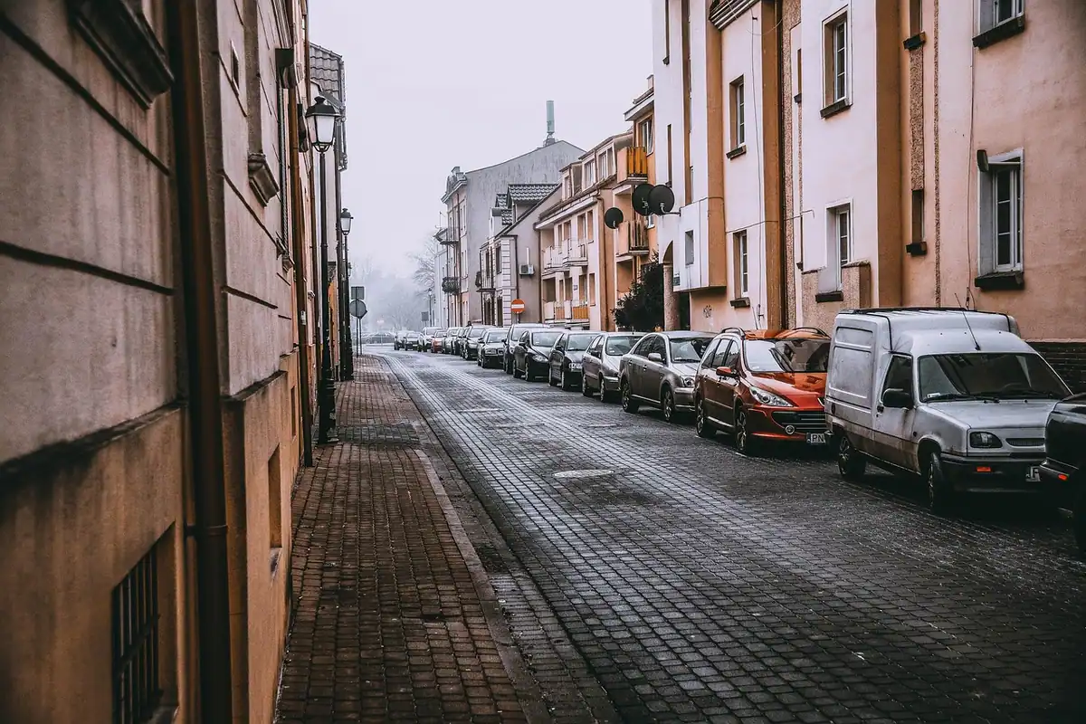 Stradă îngustă dintr-un oraș vechi cu mașini parcate lângă clădiri, fără mulți oameni
