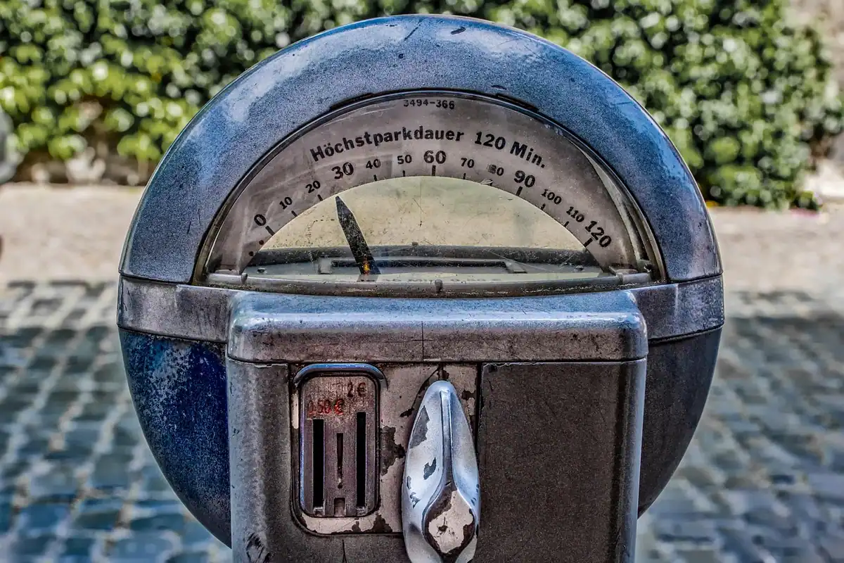 Parcometru vechi cu ceas analogic și monede, pe trotuar, lângă o stradă și tufișuri
