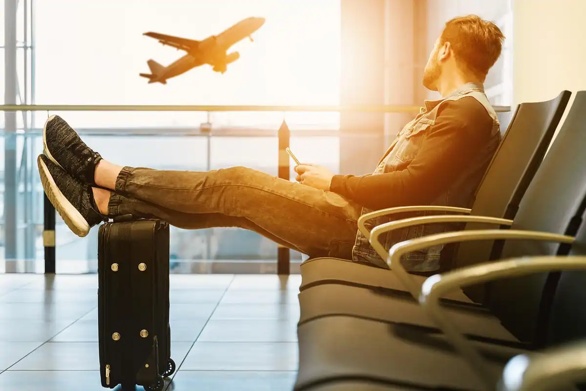 Bărbat cu picioarele pe un scaun, așteptând în aeroport, lângă o valiză mică