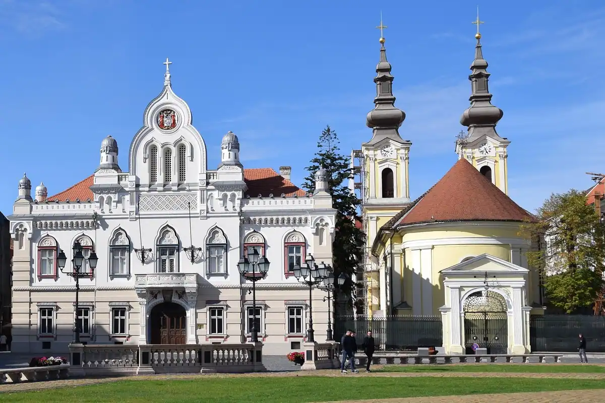 Piața din Timișoara cu biserică ortodoxă albă, turnuri și gazon verde