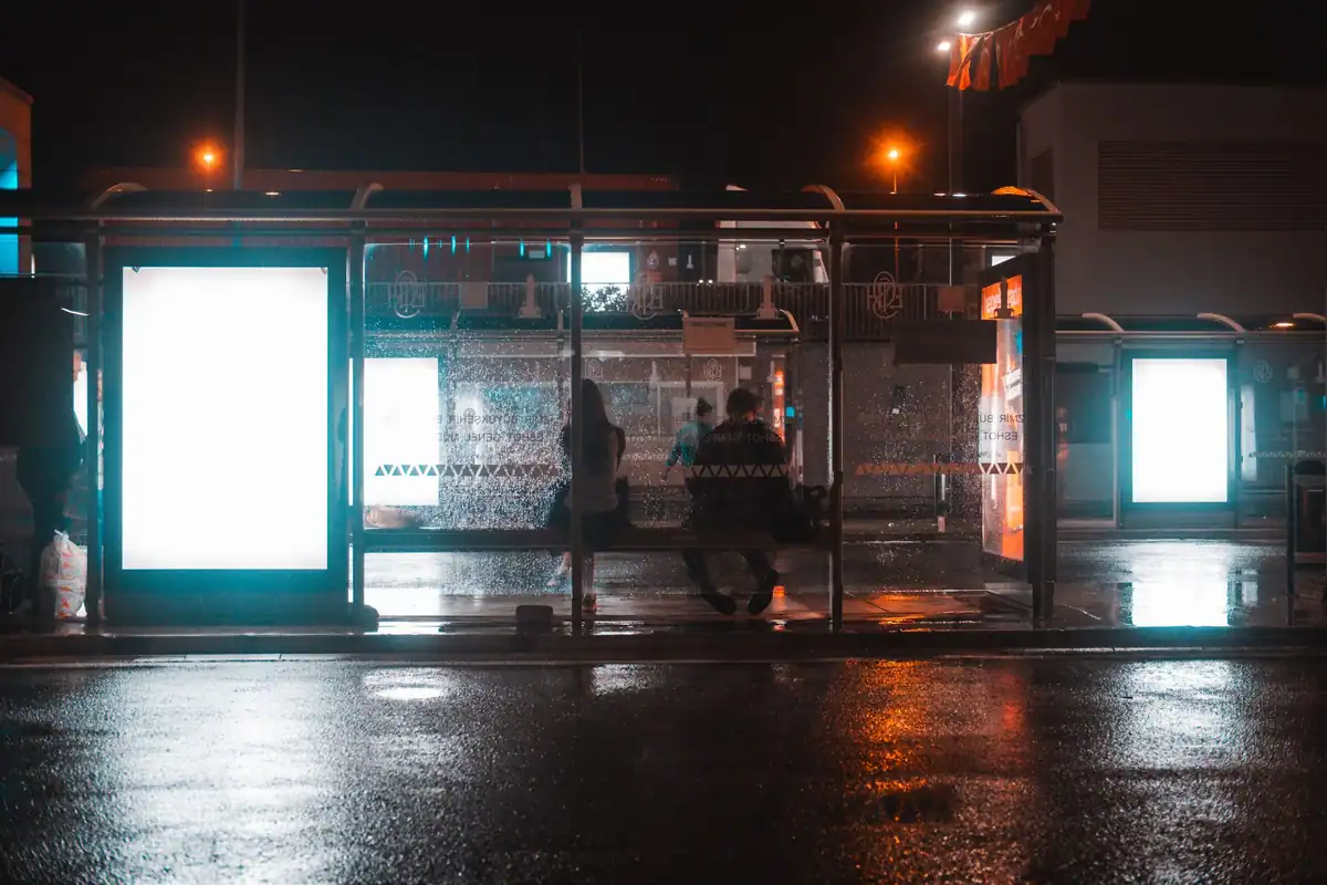 Oameni în așteptare la o stație de autobuz, noaptea, pe o stradă udă de ploaie