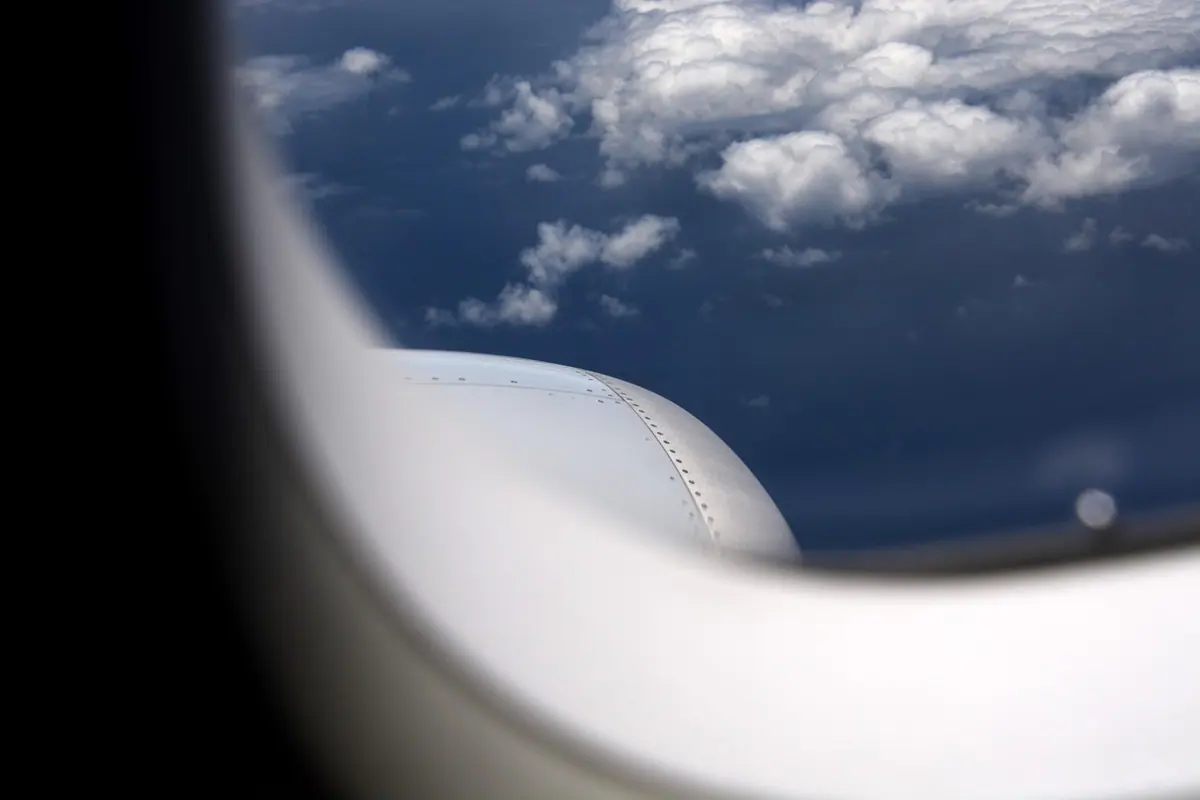 Aripă de avion văzută pe geam, cu nori albi pe cerul albastru